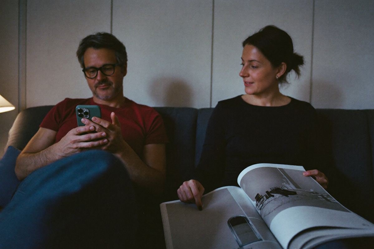 KI-Bild: Ein Paar sitzt abends auf dem Sofa. Er sucht am Handy, sie zeigt auf einen Badkatalog.